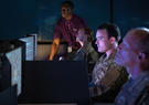 Military personnel look at computer screens as a Leidos employee assists.