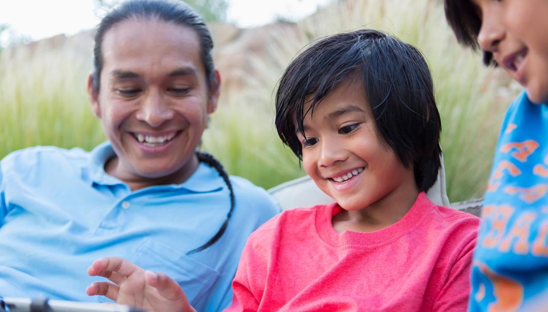 Native American father and two sons look at tablet sitting outside