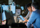 woman holding tablet next to man in front of multiple screens
