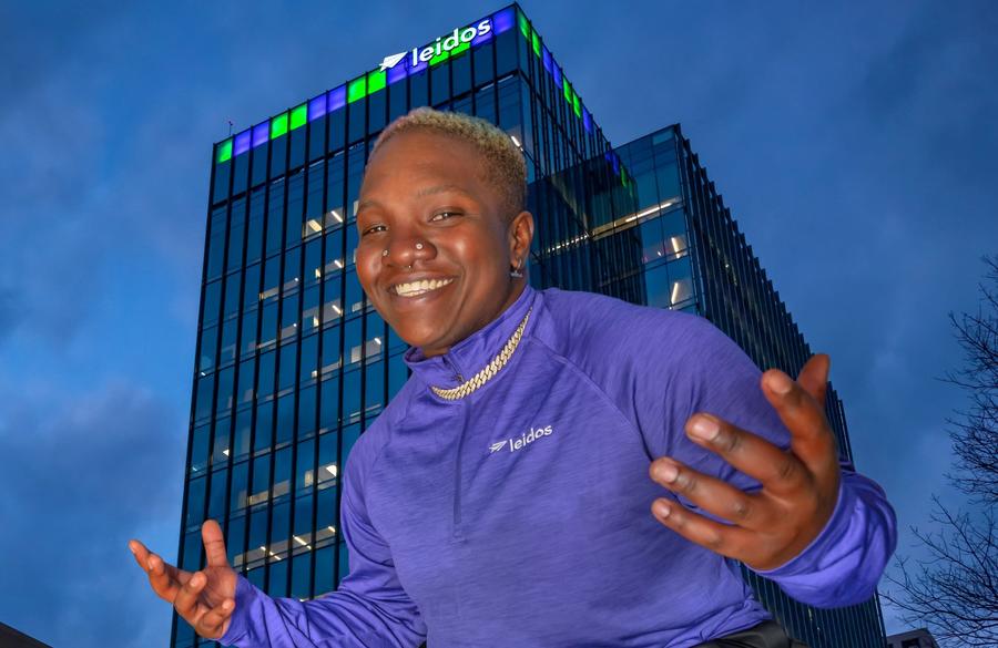 Raven Saunders posing in a Leidos pullover in front of Leidos Global Headquarters. The building is lit up in green and purple.