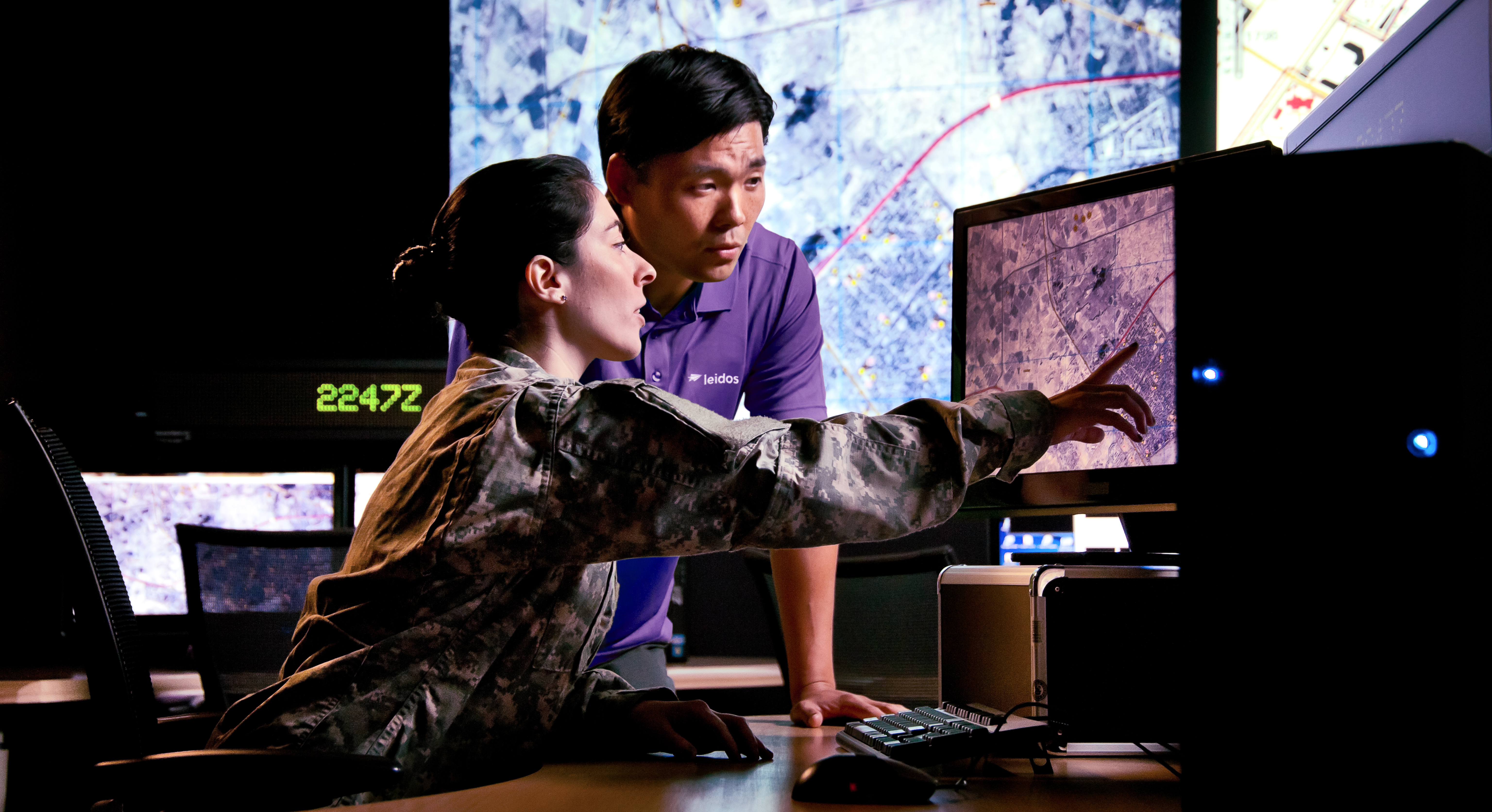 A woman in military uniform points at a screen as a man in a purple shirt looks at the screen.