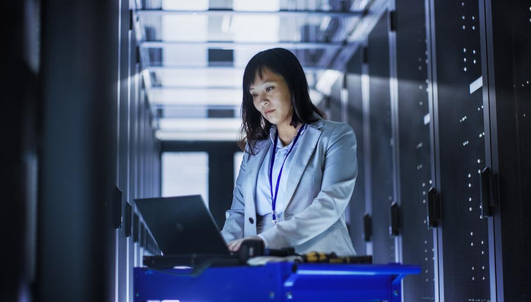 Woman in a data center standing next to server racks working on a laptop