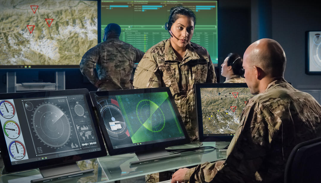 A man looking at 3 monitors in front of him while talking to another coworker