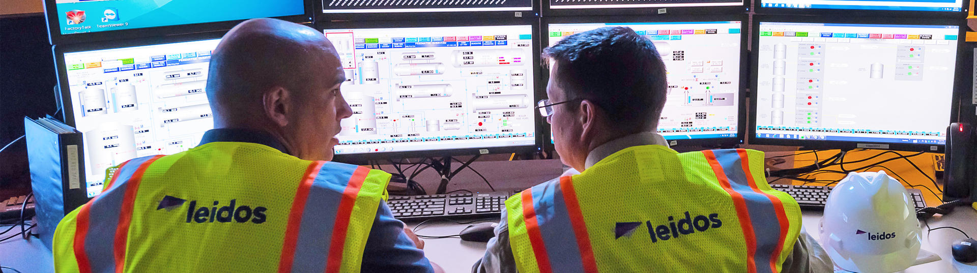 three men sitting around multiple computer monitors with safety vests on