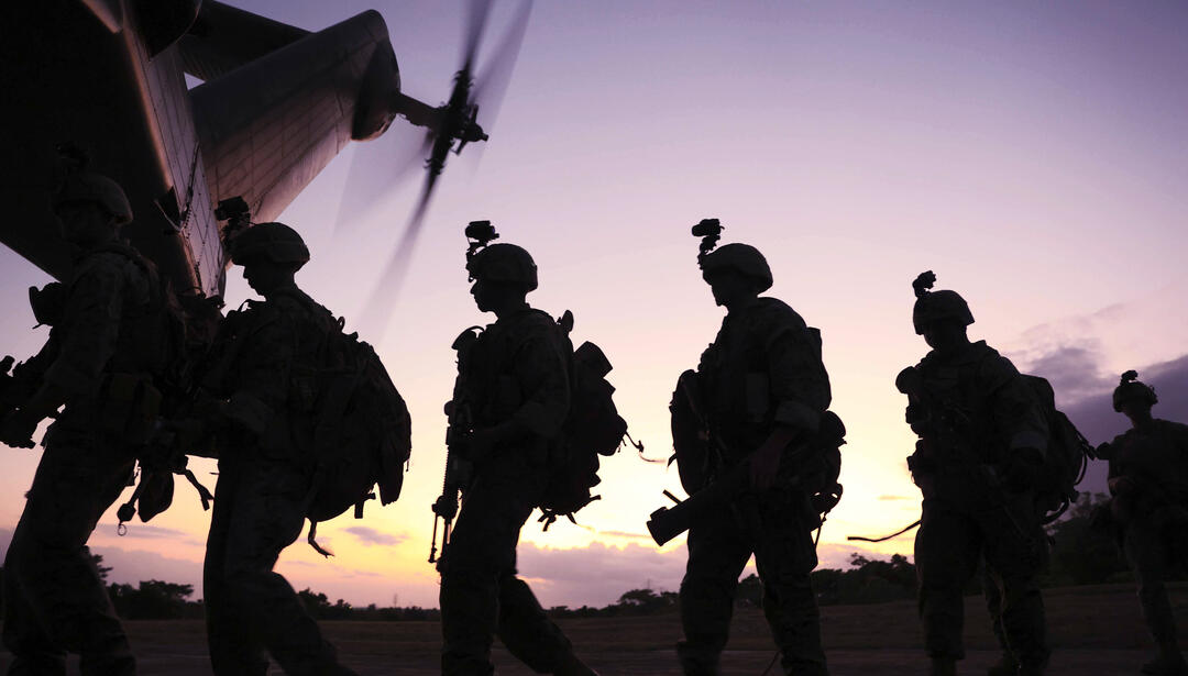Silhouetted soldiers carrying gear walk toward a helicopter at sunset, its rotor blades spinning overhead. The scene is backlit by a warm sky, emphasizing their movement and mission readiness.