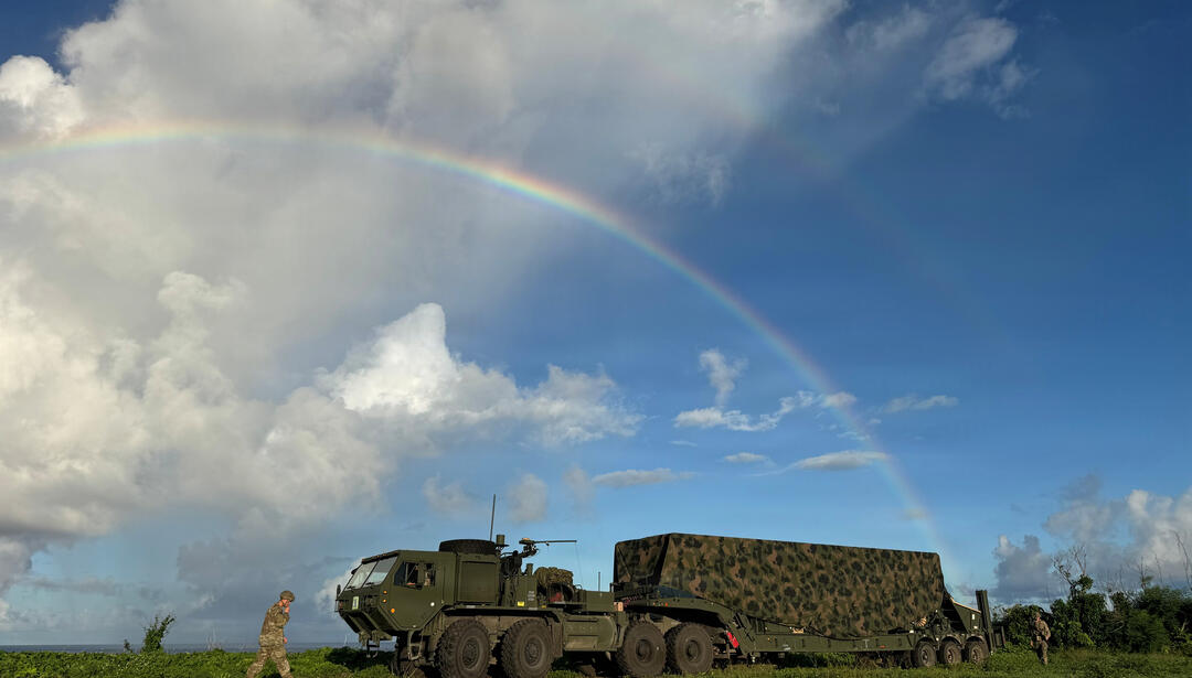A large military truck on sparse grass with a rainbow in the sky above