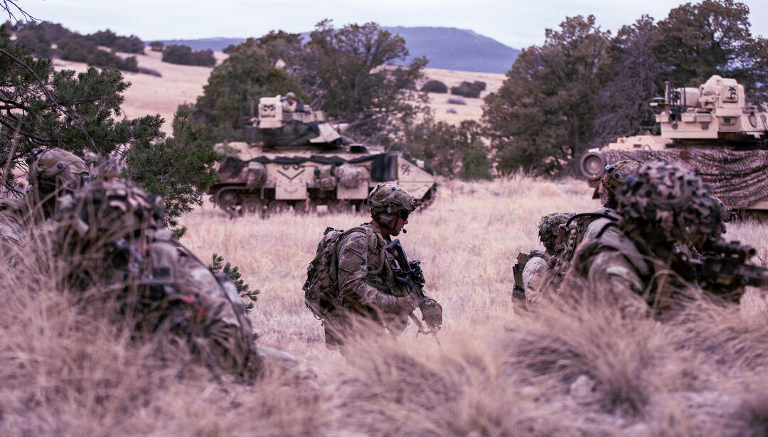 Soldiers move through rugged terrain during a military training exercise, with armored combat vehicles providing support in the background.
