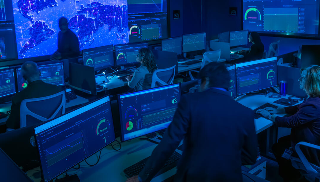 Cyber operations center with analysts monitoring multiple dashboards and global network activity across large display walls, supporting real-time cybersecurity monitoring and threat detection.