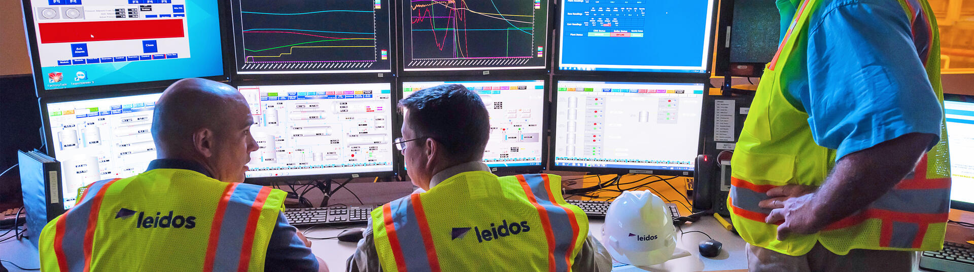 three men sitting around multiple computer monitors with safety vests on