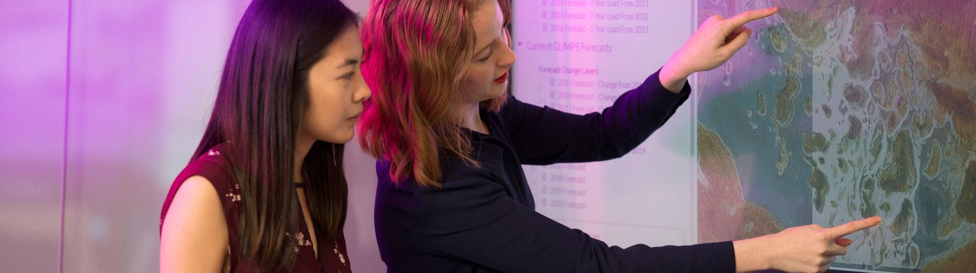 A woman looks on as another points to a map