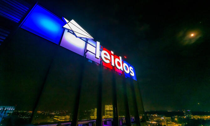 Leidos logo and wordmark pictured in a nighttime scene on top of Leidos GHQ lit up red white and blue