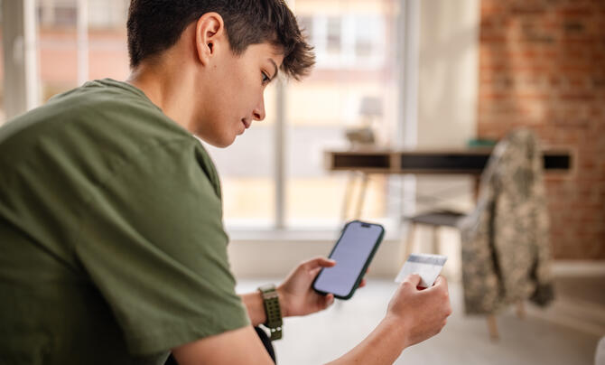 A person sitting indoors looks at a smartphone in one hand and a card in the other.
