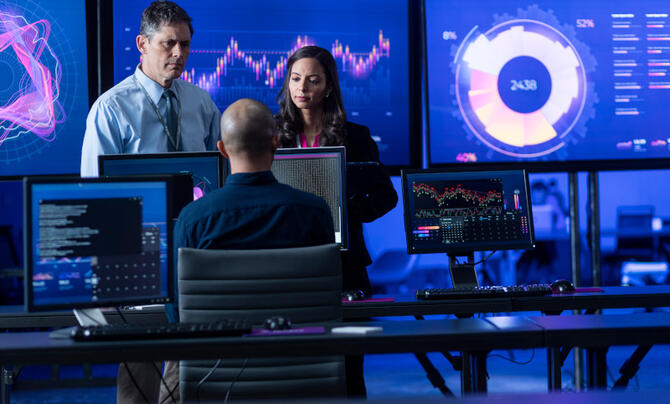 A man and a woman are standing in front of a man who is sitting at a desk with multiple computer screens, and large screens showing information and data in the background.