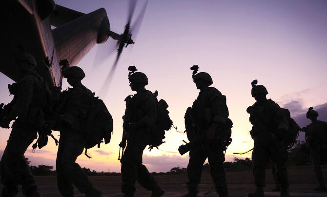 Silhouetted soldiers carrying gear walk toward a helicopter at sunset, its rotor blades spinning overhead. The scene is backlit by a warm sky, emphasizing their movement and mission readiness.