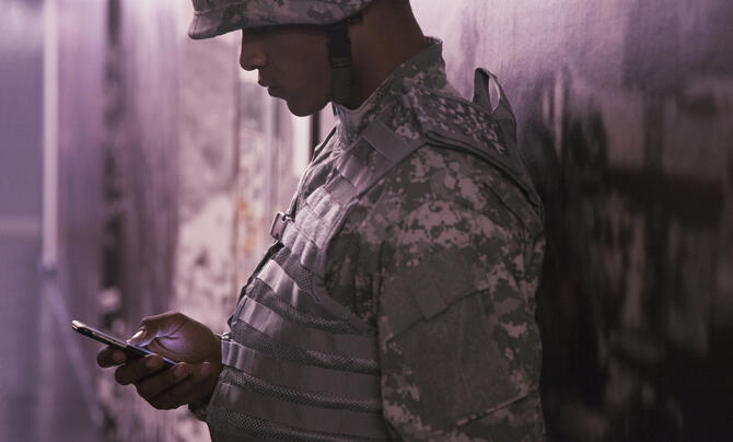 A soldier in camouflage uniform and helmet leans against a wall, looking down at a smartphone. The scene is softly lit with a muted, slightly somber tone.
