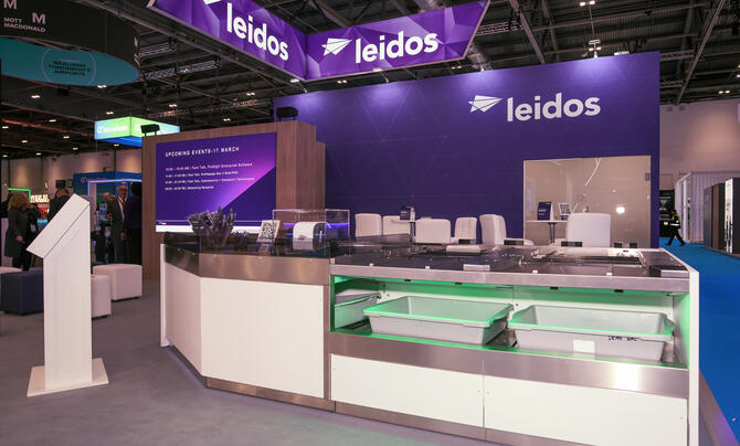 A trade show booth with prominent “Leidos” branding features a modern display counter and seating area. Overhead signage and screens highlight the company’s presence in a large exhibition hall.