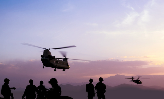 Six soldiers in silhouette standing on top of a large hill or mountain facing the sunset while a Chinook and other unidentified helicopter fly past.