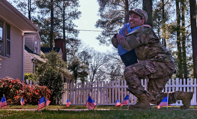 A male soldier bends on one knee to hug a child in front of a home along a path with green grass and small U.S. flags along the path.