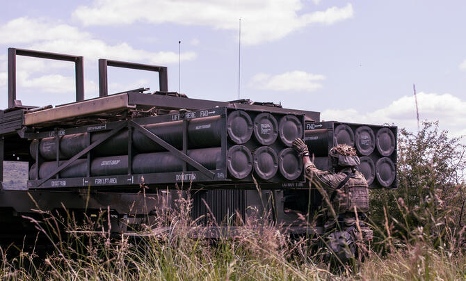 A soldier in tactical gear inspects a truck-mounted system loaded with cylindrical equipment in a grassy field under a partly cloudy sky.