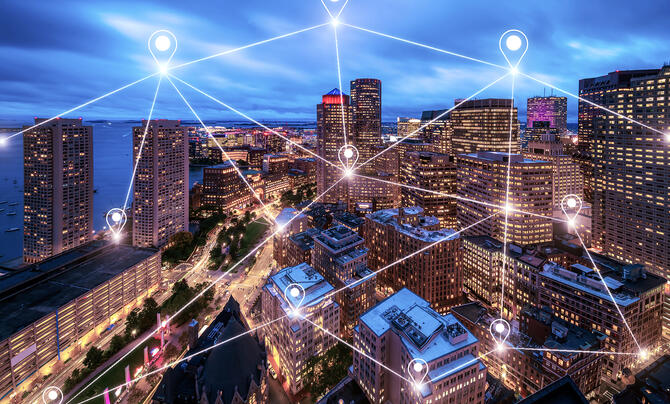 An aerial view of a city skyline at dusk features glowing location markers and connected lines overlaying the buildings, illustrating a digital network across an urban environment.