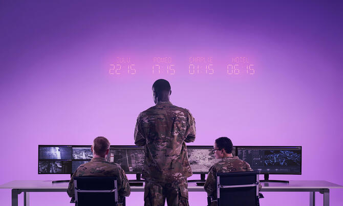 Military personnel monitor surveillance and operational data on multiple screens in a command center illuminated by purple lighting.