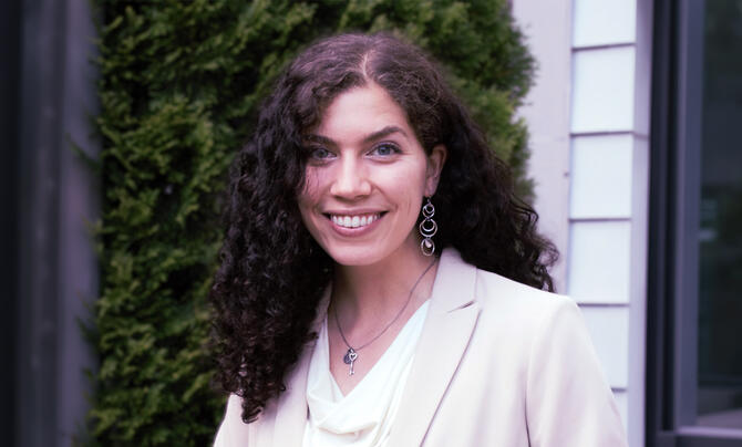 A smiling woman with curly dark hair stands outdoors wearing a light-colored blazer and earrings, with greenery in the background.