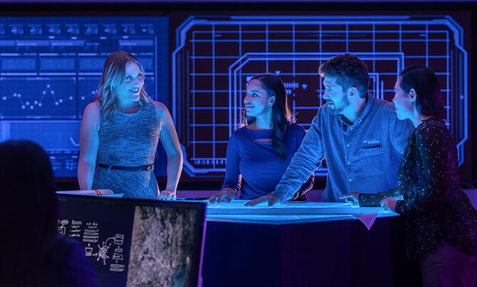 A team of professionals collaborates around a digital workspace, reviewing data and plans in a technology operations center illuminated by large interactive displays.