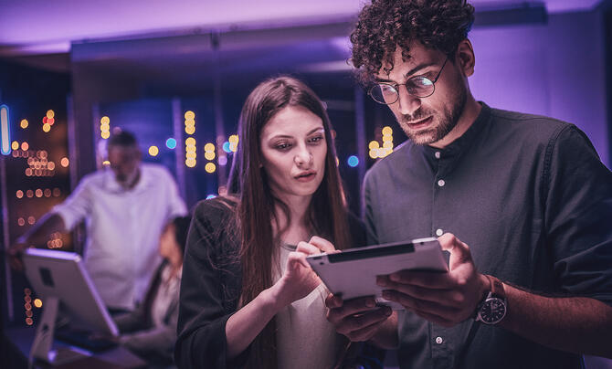 Two professionals collaborate over a tablet in a technology-focused workspace, reviewing information together under ambient purple lighting.
