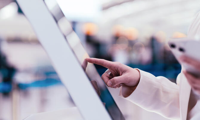 Close-up of a traveler using a touchscreen self-service kiosk while holding a smartphone, illustrating digital identity verification and contactless passenger processing at an airport.