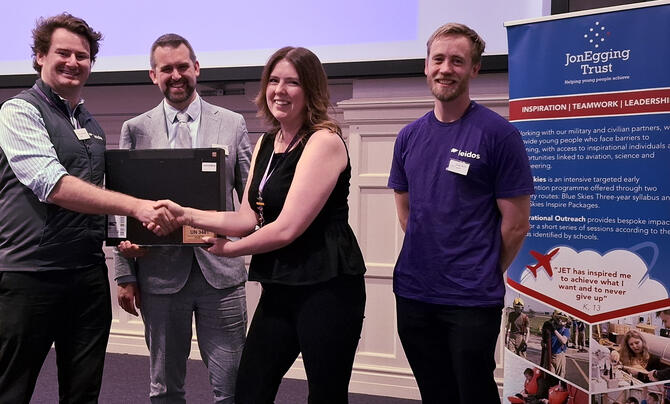 Leidos employees present a donated laptop to representatives of the Jon Egging Trust during a community partnership event, standing beside a Jon Egging Trust display banner.