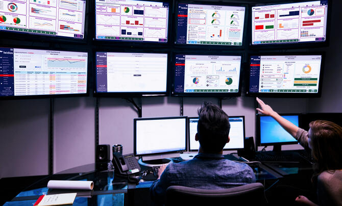 Two professionals monitor operational dashboards on multiple large displays, reviewing performance metrics, analytics, and system status in a technology operations center.