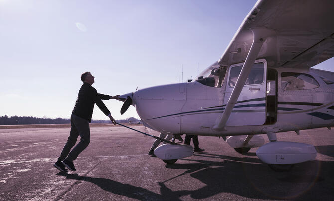 A person pushes a small single-engine aircraft across an airport ramp before flight, with another individual boarding through the open cabin door under a clear sky.