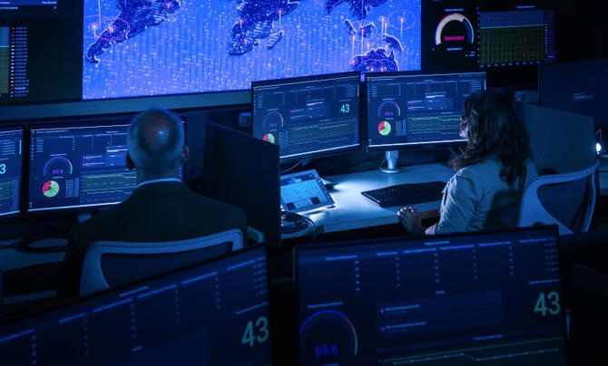 A dark operations control command center, with two people sitting at desks viewing computer screens.