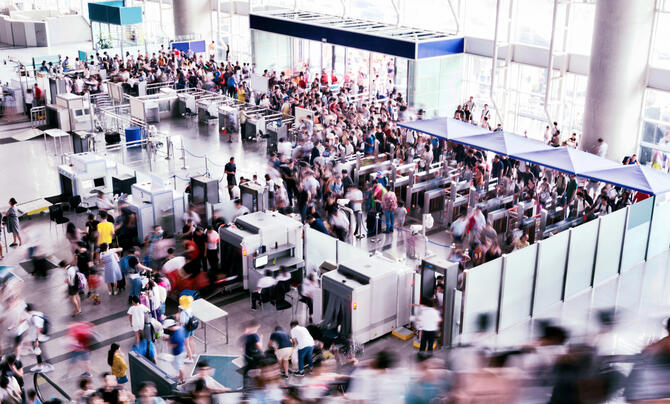 Crowds of travelers move through security screening checkpoints and queues inside a busy airport terminal.
