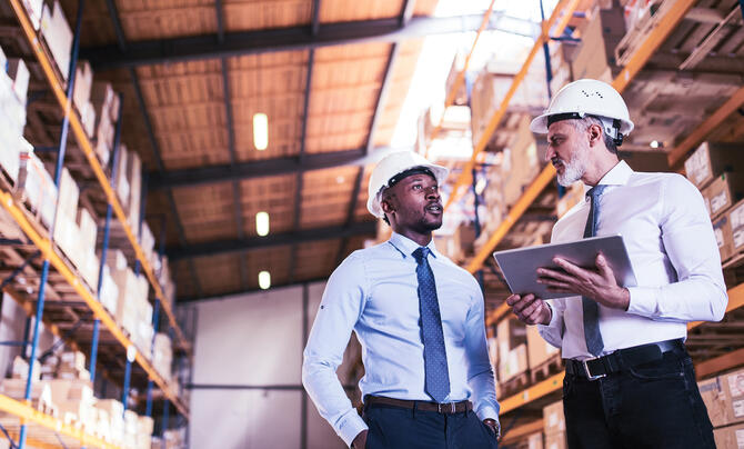 Two professionals wearing hard hats discuss operations while reviewing information on a tablet in a warehouse.