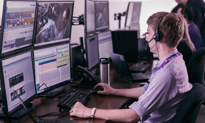 Leidos employee wearing a headset monitors multiple computer screens displaying operational data and video feeds.