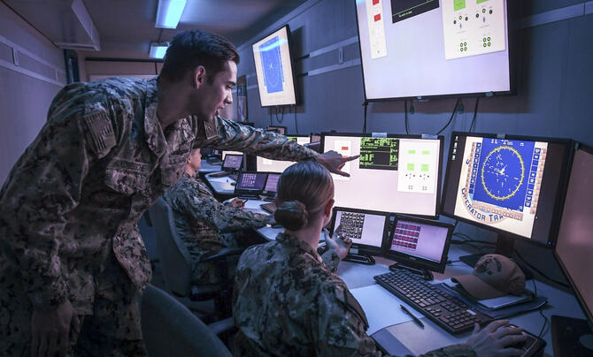 Two military servicemembers in a small computer operations room viewing and pointing at computer screens showing simulated data and maps 