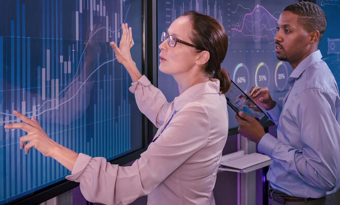 Two professionals analyze data visualizations on large digital displays, with one pointing to a chart while the other holds a tablet.