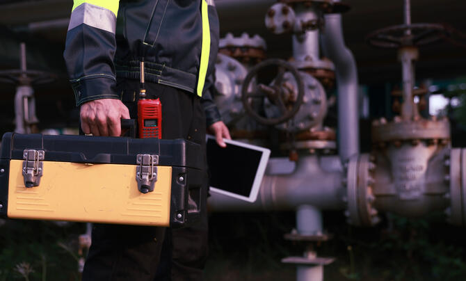 Industrial worker in high-visibility clothing carries a toolbox and tablet beside large pipes and valves at an industrial facility.