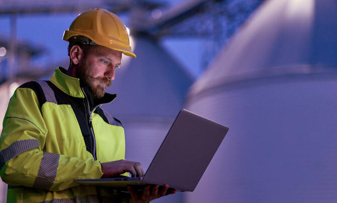 Engineer wearing a yellow hard hat and high-visibility jacket uses a laptop inside an industrial facility.