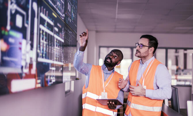 Two engineers wearing orange high-visibility vests review operational data on large digital displays, with one pointing at the screen while holding a tablet.