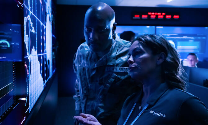 U.S. Air Force service member and Leidos employee analyze data on large digital displays in a military cyber operations center.