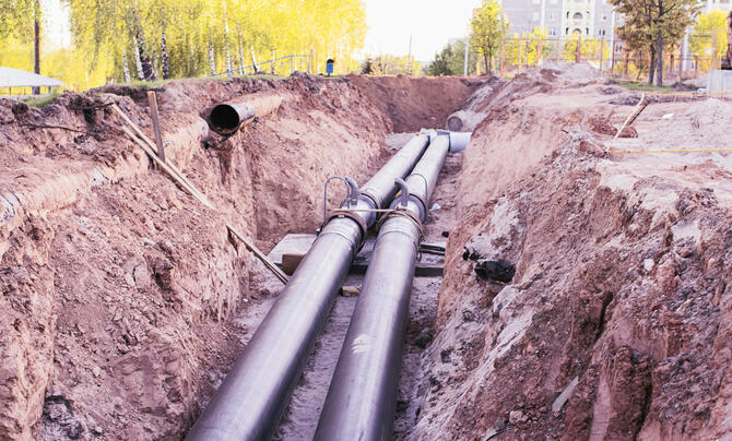 Two large metal pipelines run through an excavated trench at a construction site, surrounded by earth and construction materials.