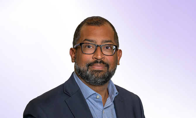 A headshot style photo of Stephen Vather. He has dark-rimmed glasses and is wearing an open collar blue dress shirt with a gray jacket. He has a closely trimmed beard.