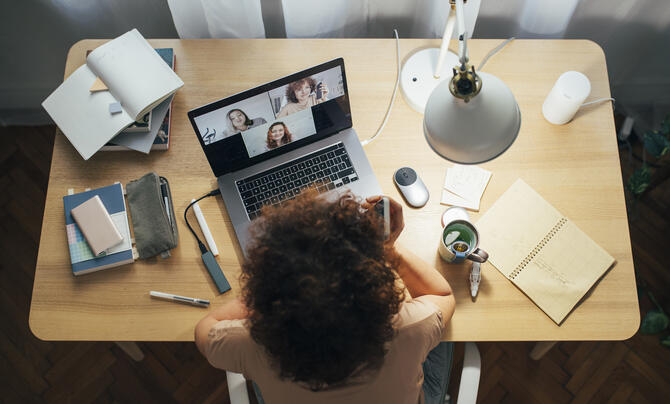 A woman working at home with a laptop open on a desk, surrounded by notebooks and pens.