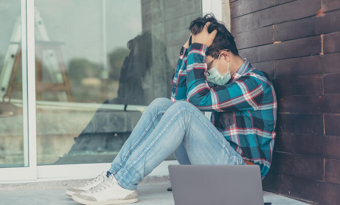 Man wearing mask with head in his hands with laptop next to him