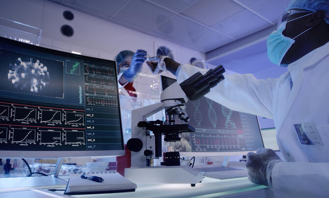Two masked surgeons in a lab pass samples to each other across a table which holds a microscope and computer monitors
