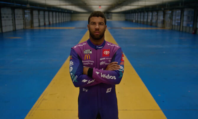 NASCAR Driver Bubba Wallace in Leidos racing suit standing in an empty garage with arms crossed