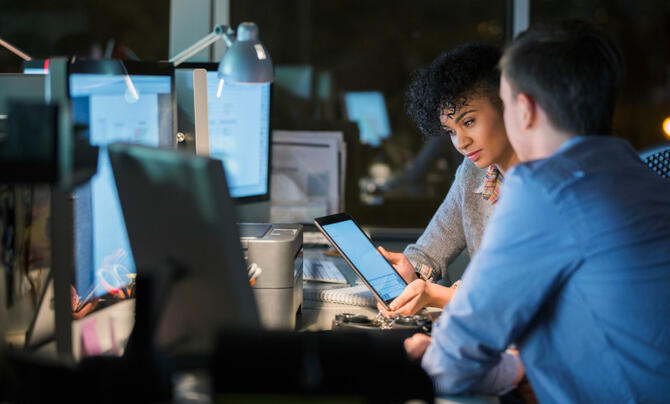 woman holding tablet next to man in front of multiple screens