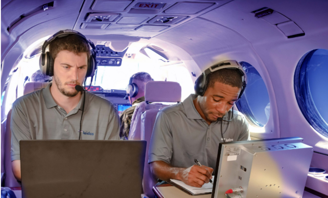 Two men in an airplane wearing headsets working on computers